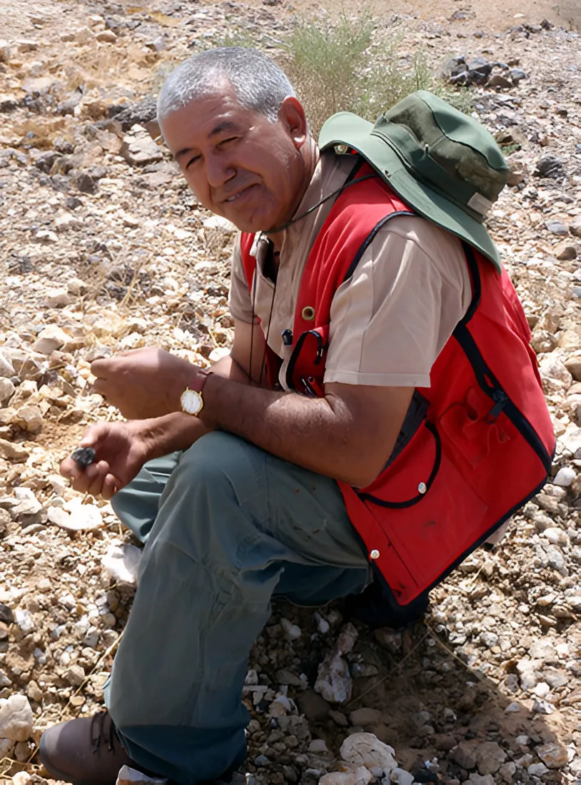 Man in a red vest and sun hat crouching on stony ground