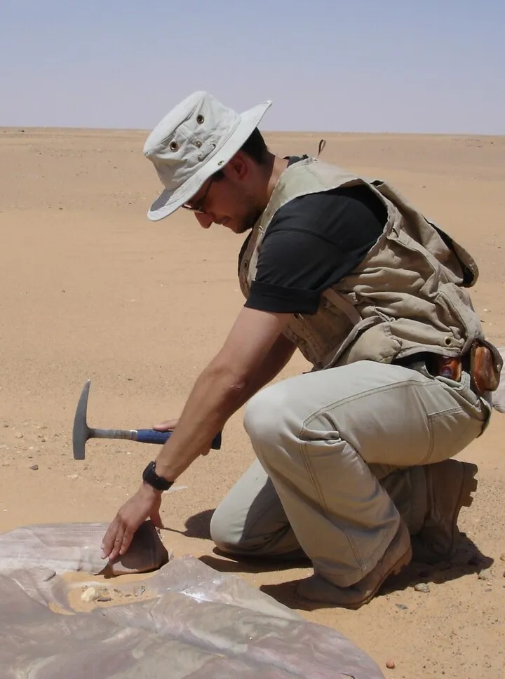 Geologist in a field hat crouching on desert sand with a rock hammer