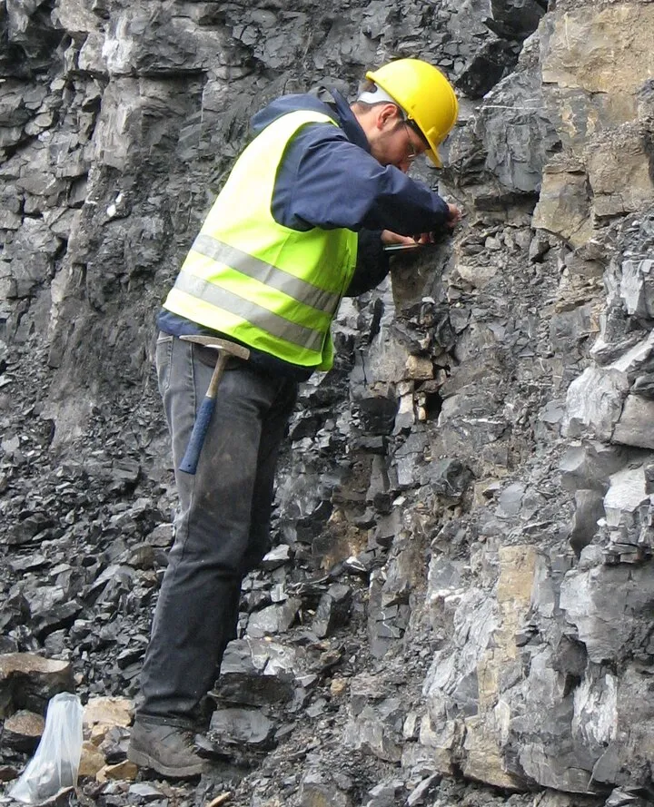 Geologist in a hard hat and high-visibility vest examining a dark rock face