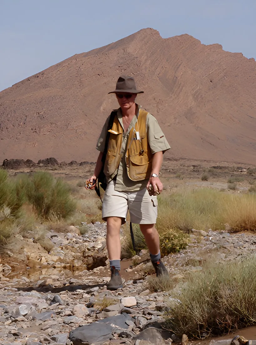 Man in a hat and vest walking along a dry streambed below a mountain