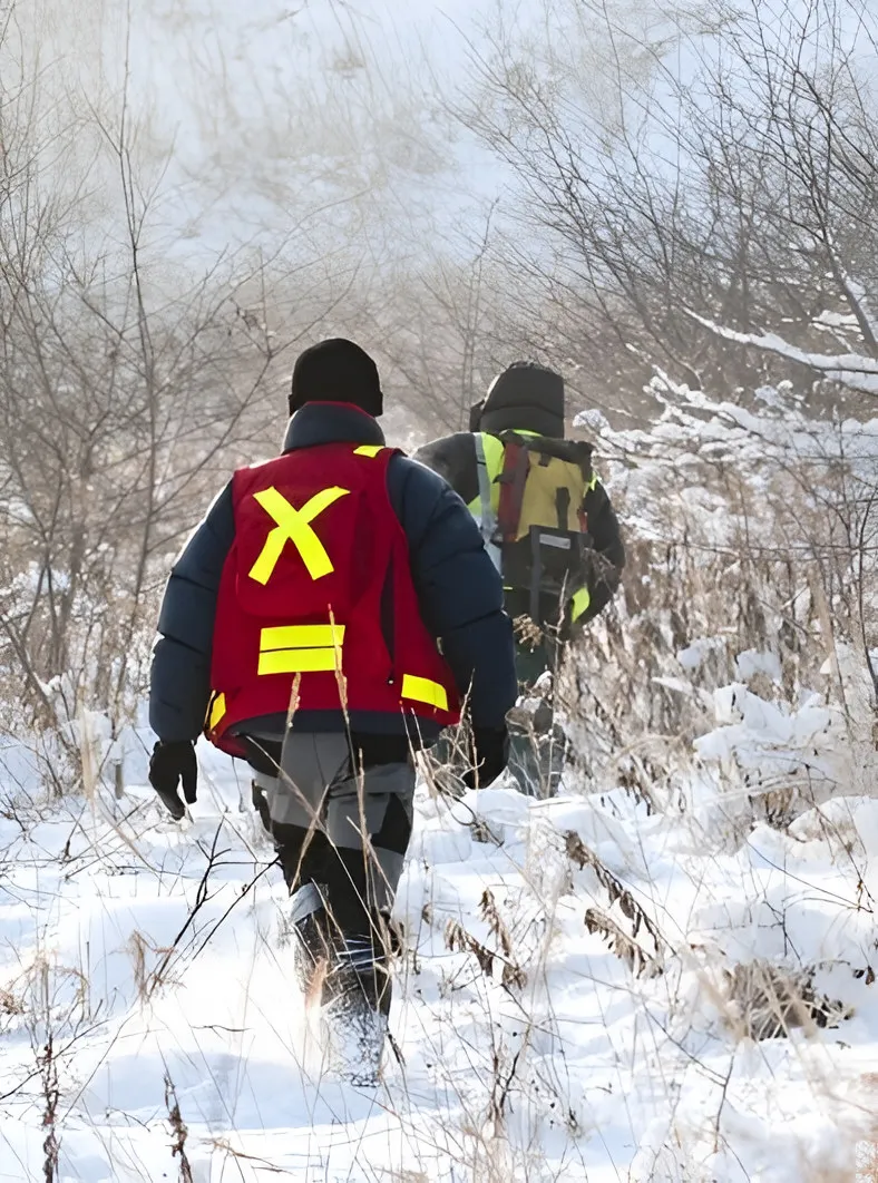 Two people in high-visibility vests walking through snowy brush